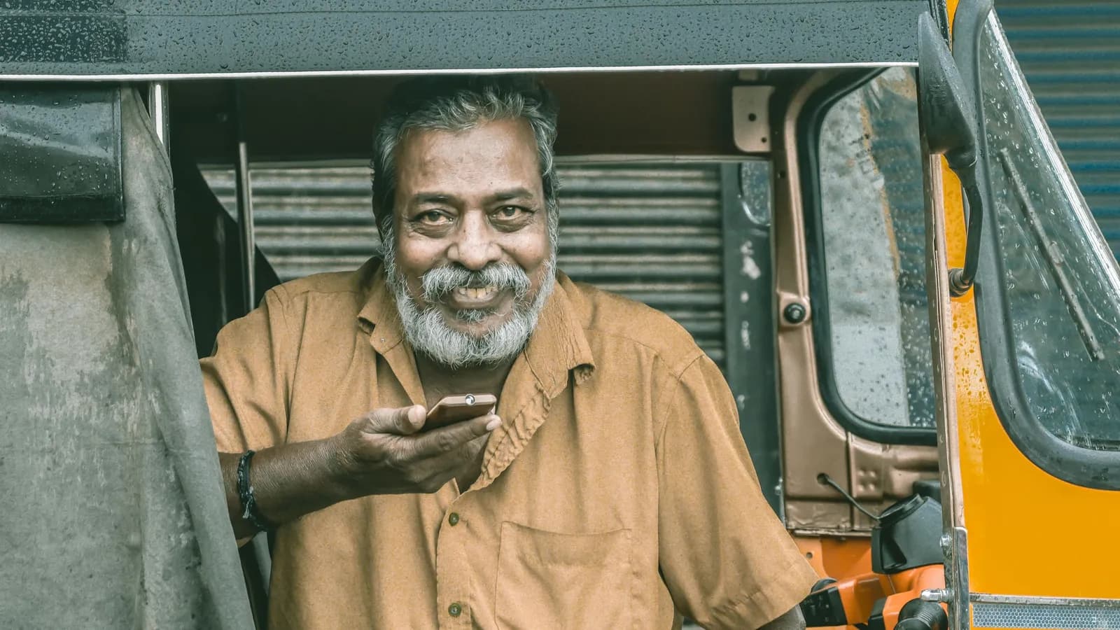 നരച്ച താടിയുള്ള ഒരു മുതിർന്നയാൾ ഓട്ടോറിക്ഷയിലിരുന്ന് ഫോൺ കയ്യിൽ പിടിച്ച് ചിരിക്കുന്നു.
