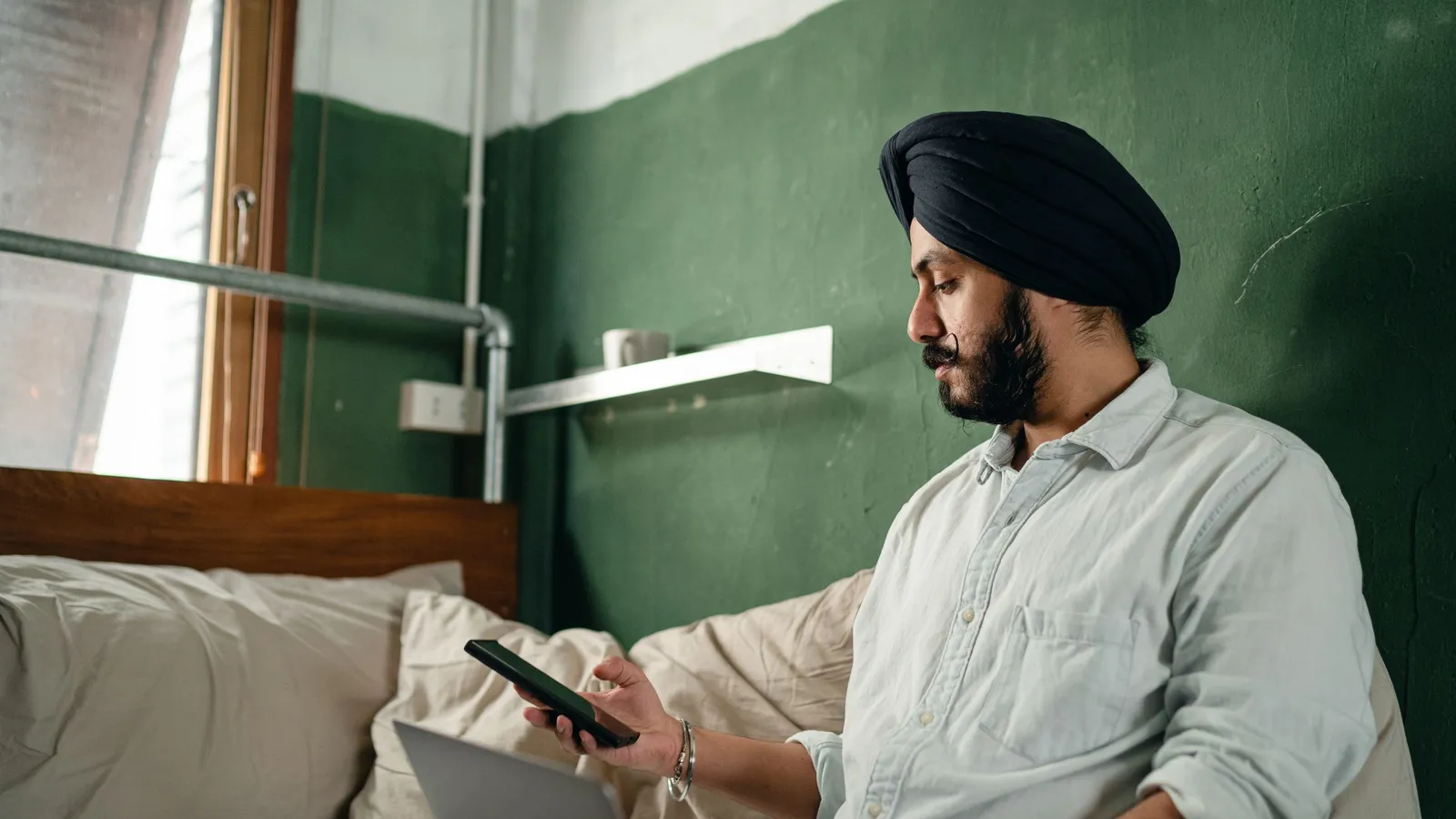 A man in a turban texts on his phone with a laptop open beside him.