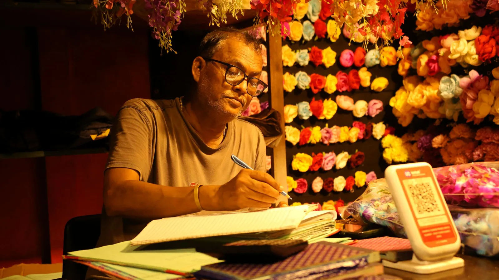 A shopkeeper writes in a ledger at his counter, with a QR code standee beside him and flowers behind.