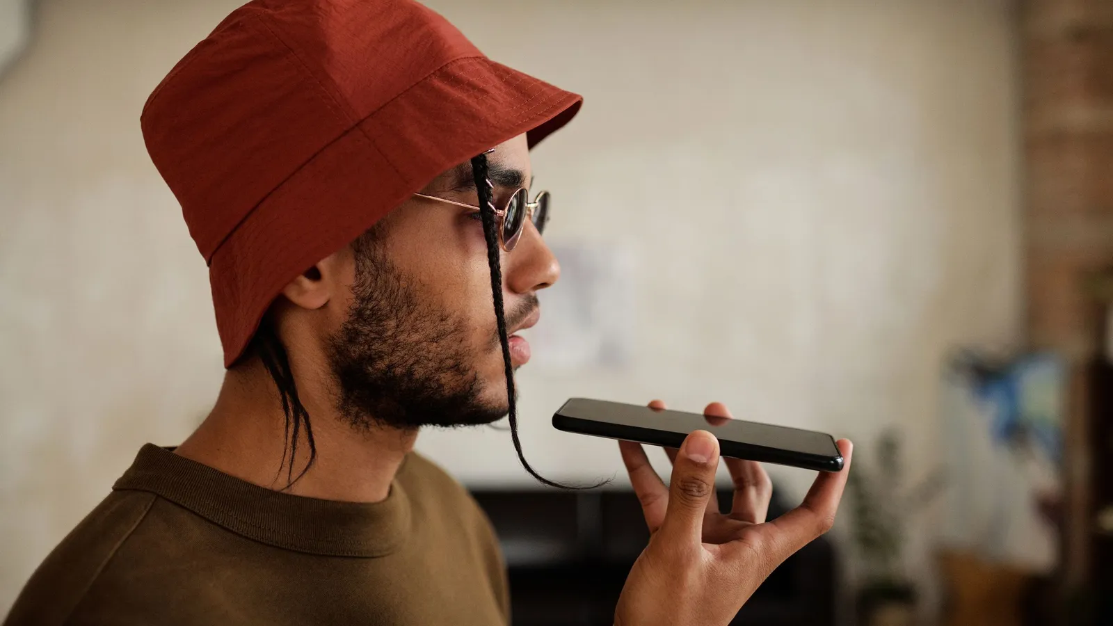 A young man in a red bucket hat speaks into his smartphone held up in front of him at home.