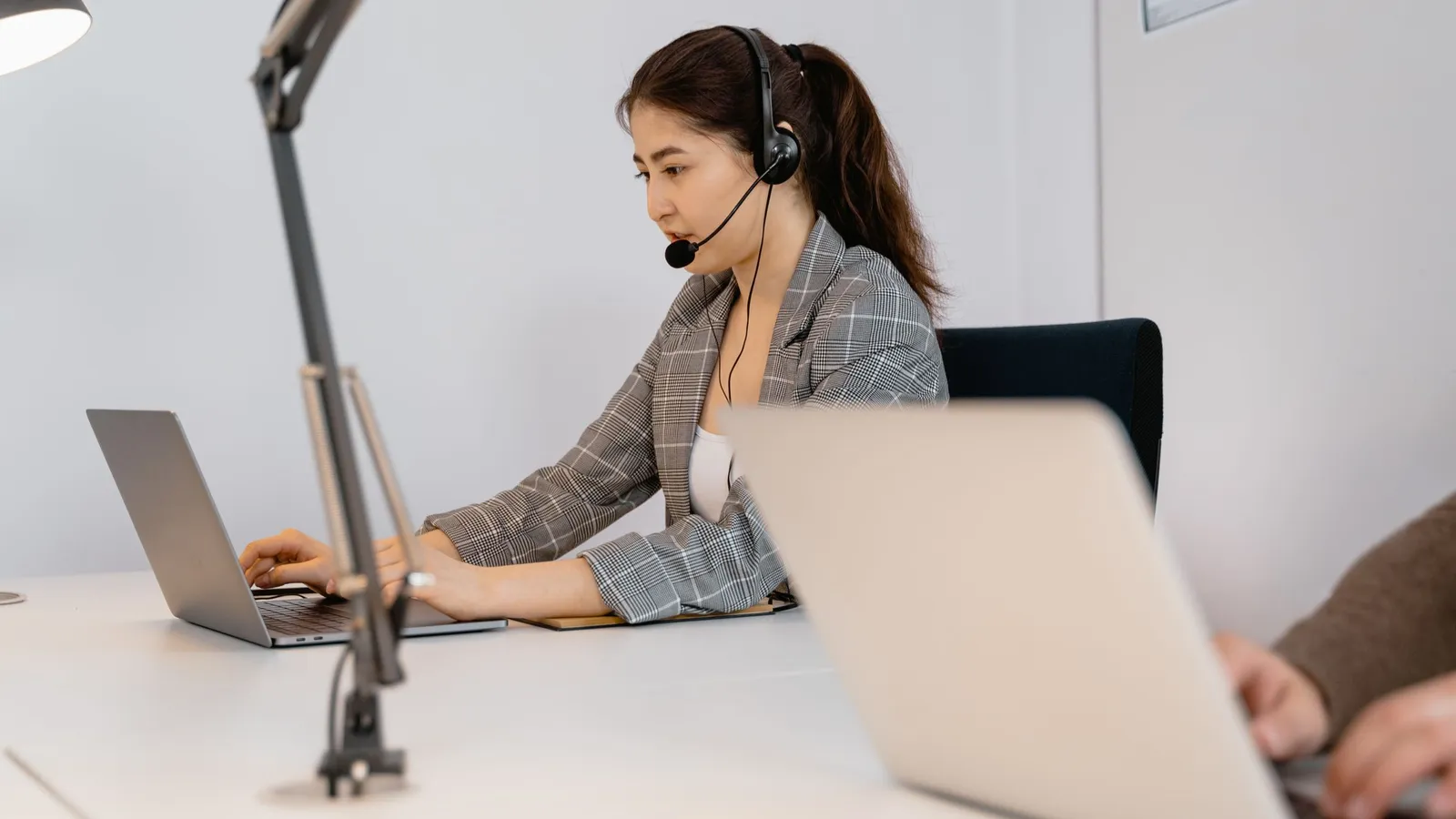 A woman wearing a headset with a microphone speaks while working at a laptop on a desk.