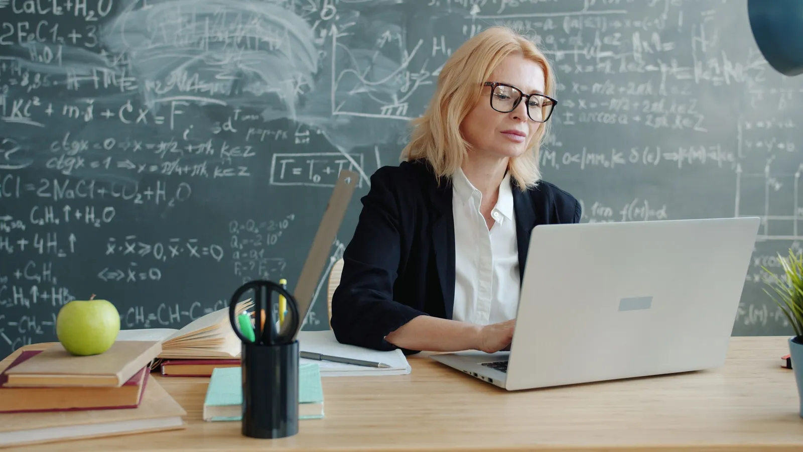 A teacher works on a laptop at her desk in front of a chalkboard covered in equations.