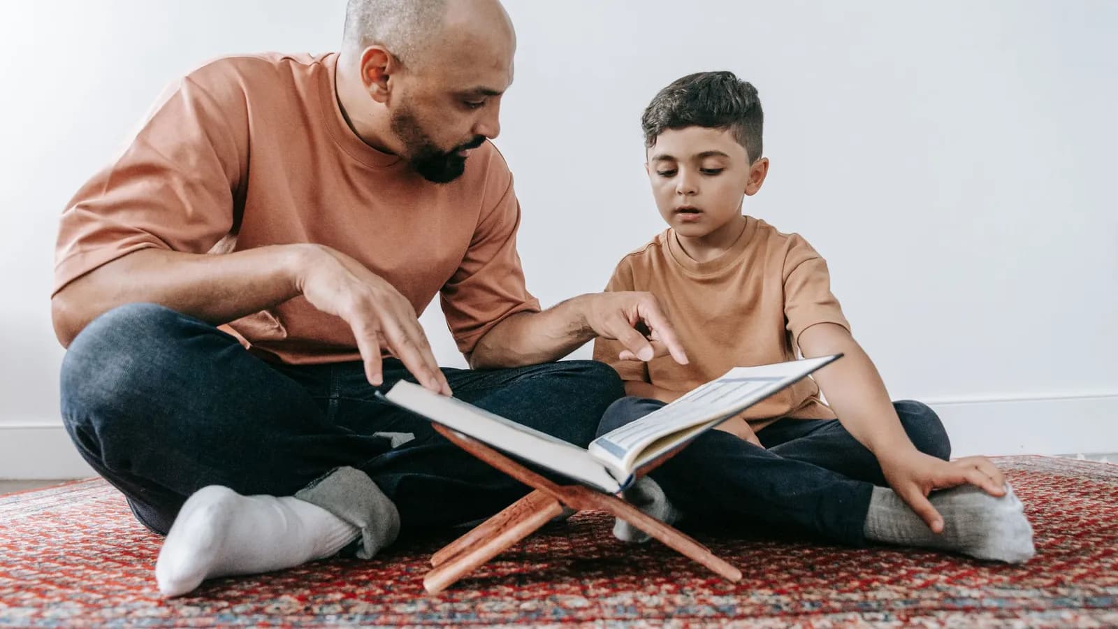 A father sits on the floor with his young son, pointing at words in a book.