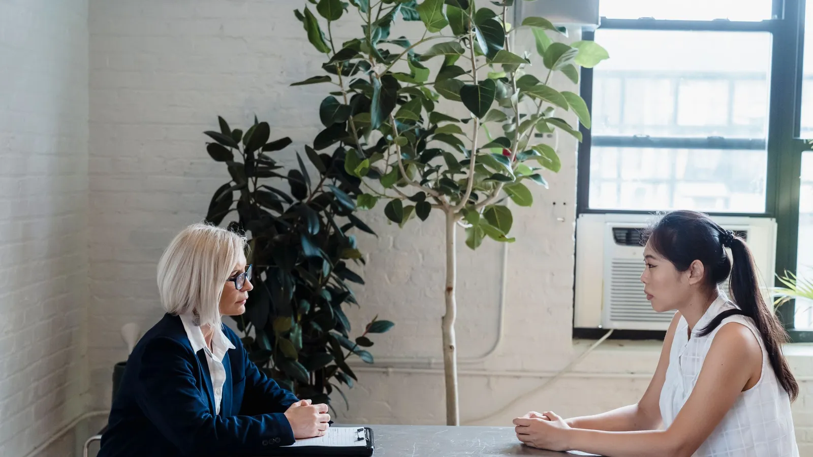 A young woman answers questions from an examiner seated across a table from her.