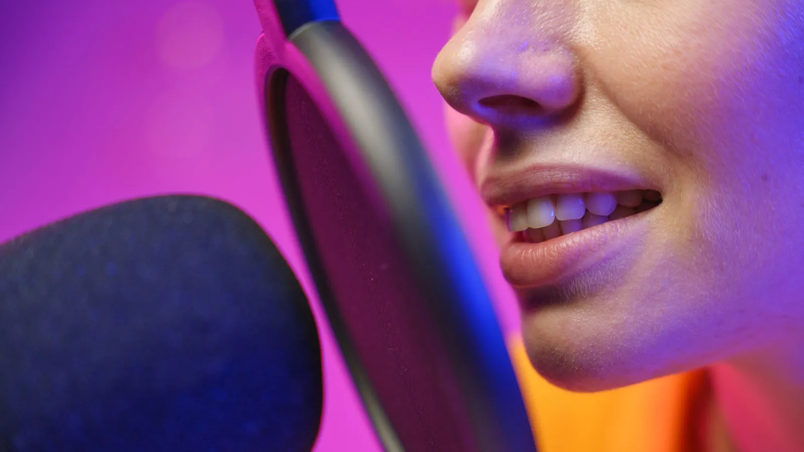 A close-up of a woman's mouth speaking into a microphone with a pop filter under purple light.