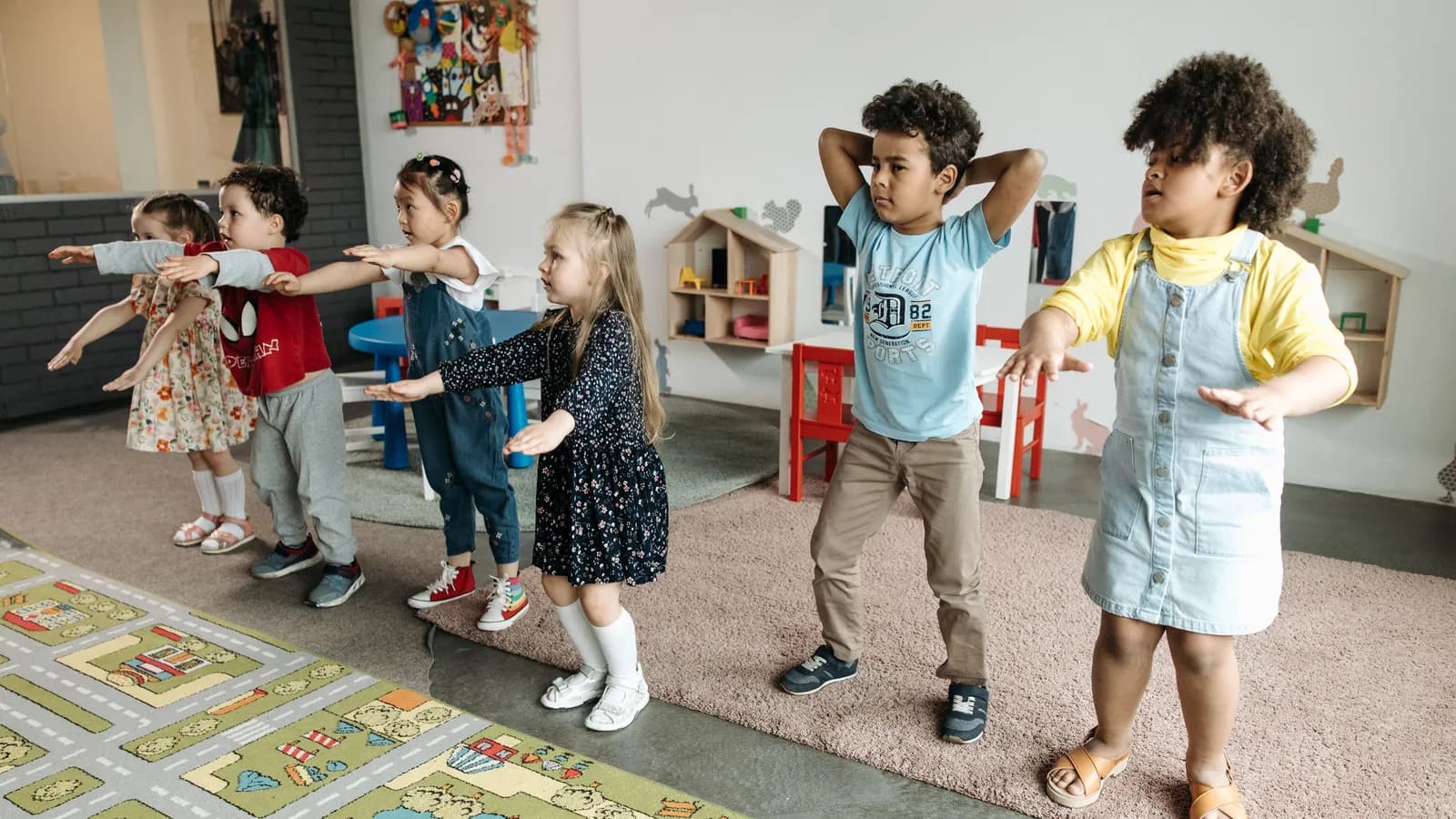 Young children stand in a classroom stretching out their arms, copying an action.