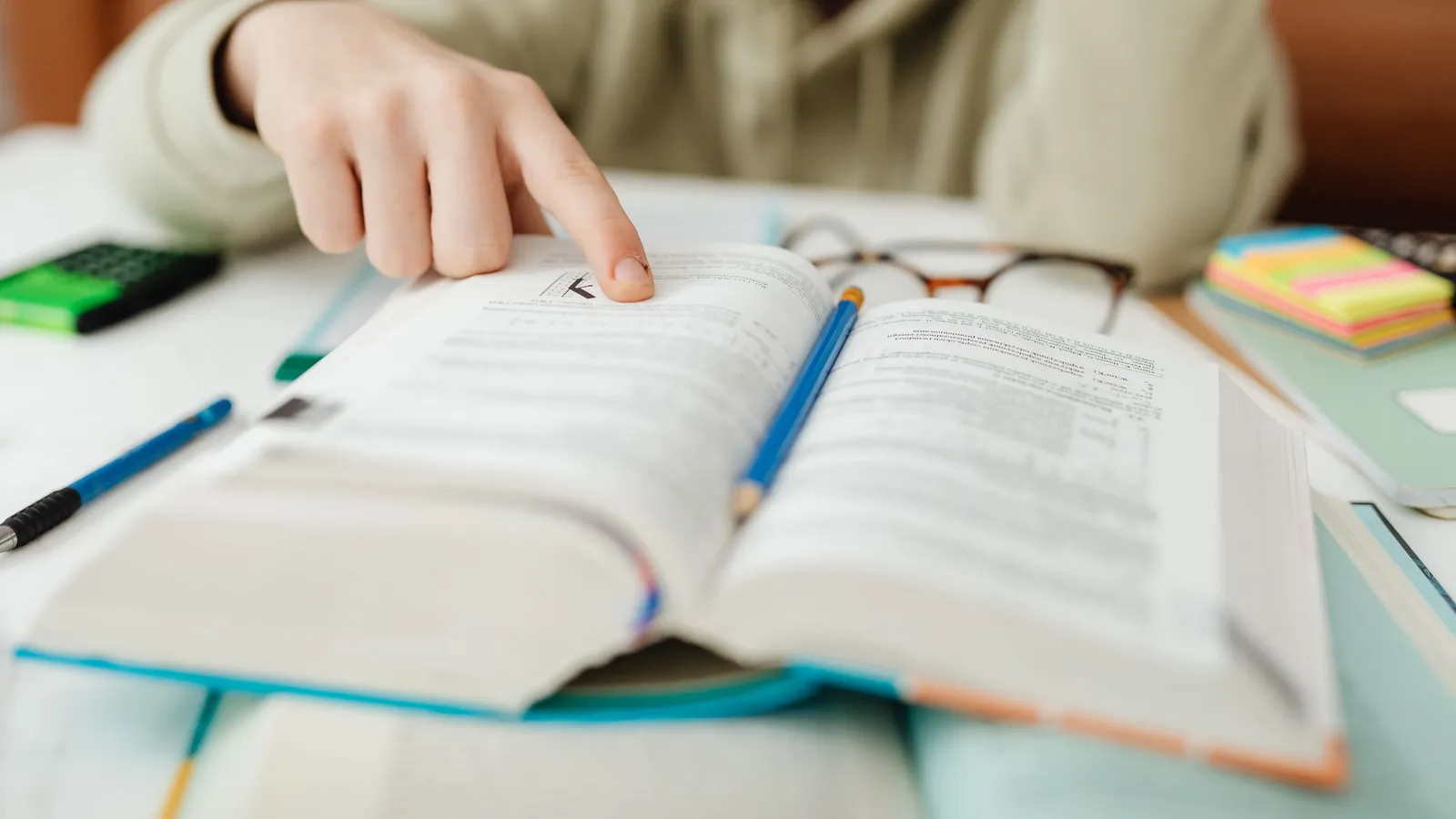 A student points to a line in an open textbook, a pencil and glasses beside it, while preparing for an exam.